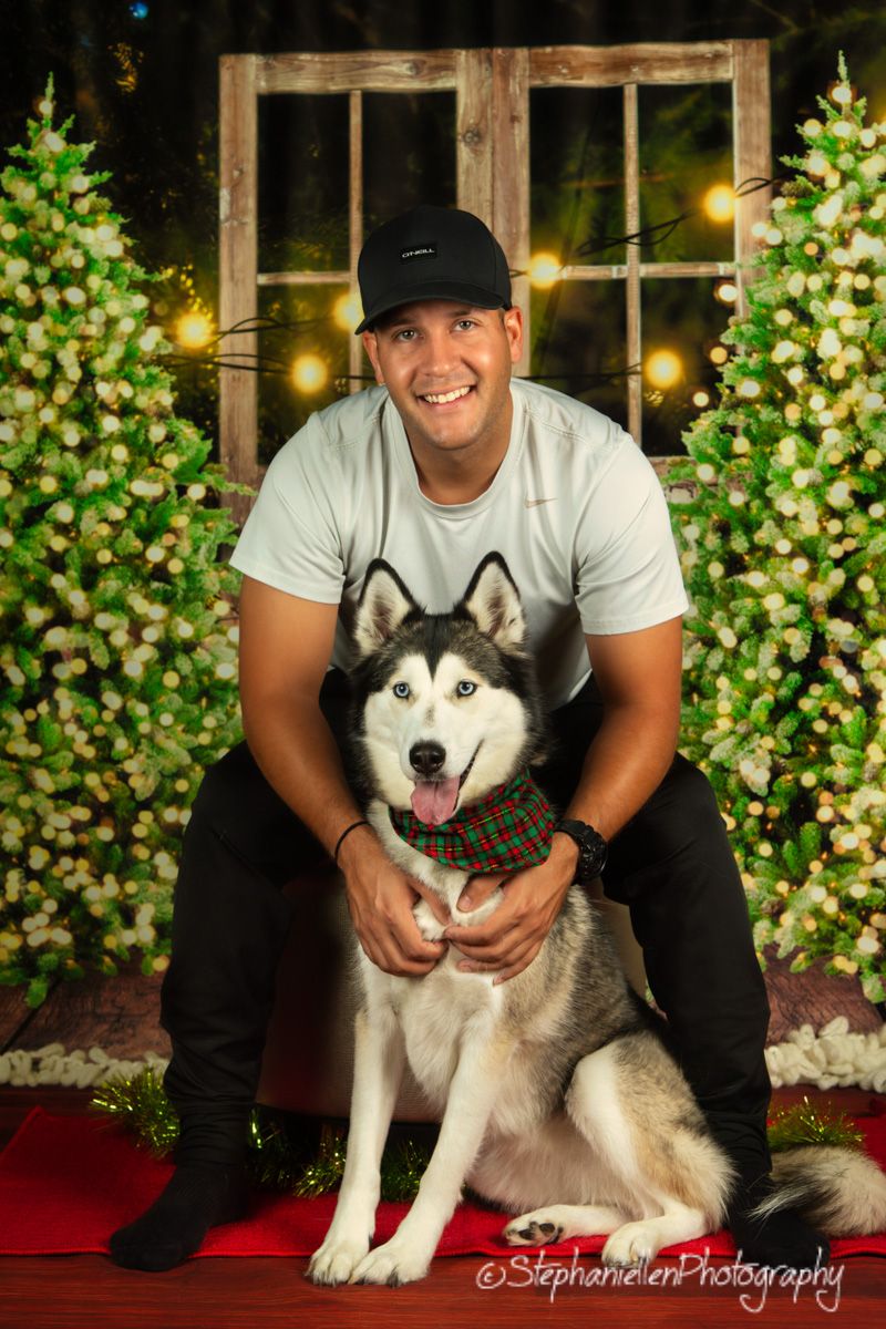 Smiling man in cap posing with a Siberian Husky wearing a plaid scarf beside Christmas trees.