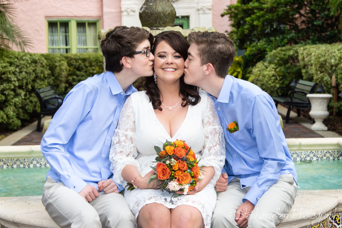 Bride in white lace dress receives kisses from two boys beside garden fountain.