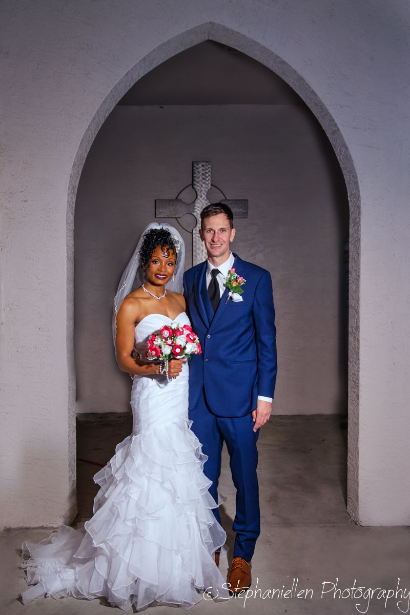 Interracial bride and groom posing together under a stone arch with a cross at their wedding ceremony.
