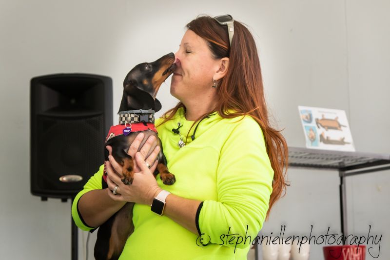 Woman in neon yellow shirt kisses small dachshund dog wearing red bandana while holding it indoors.