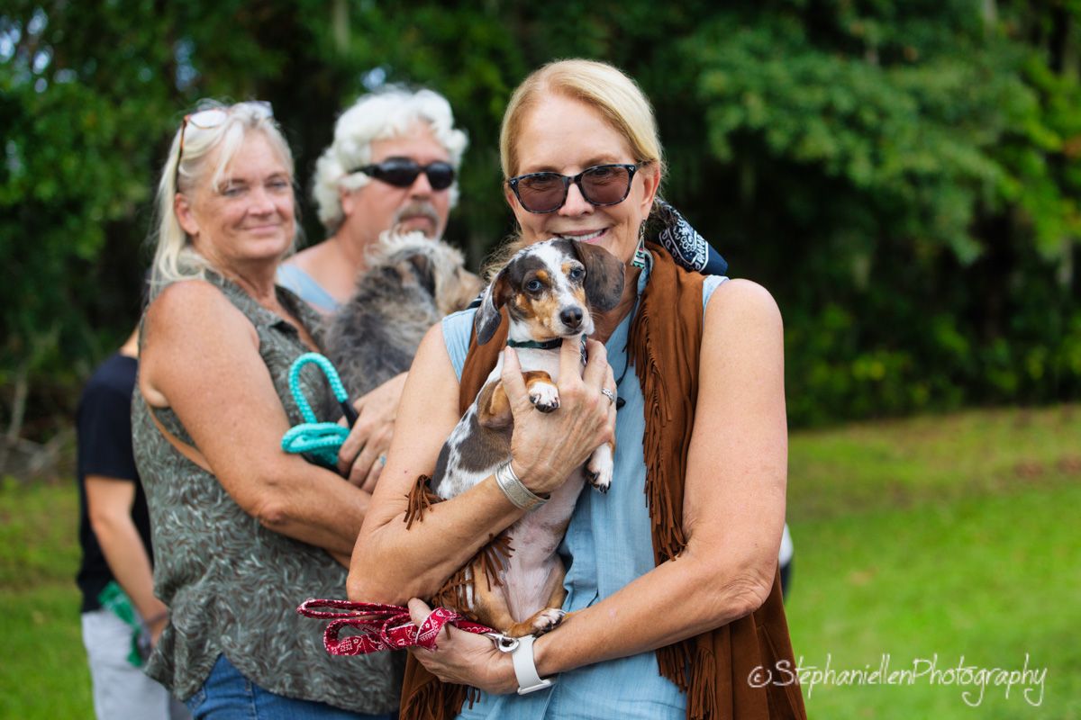 Two women holding small dogs outdoors at a casual gathering, with a man standing behind them among green trees.