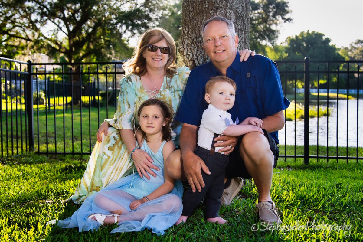 Family of four poses by a tree in a sunny green park, with parents and two young children smiling outdoors.