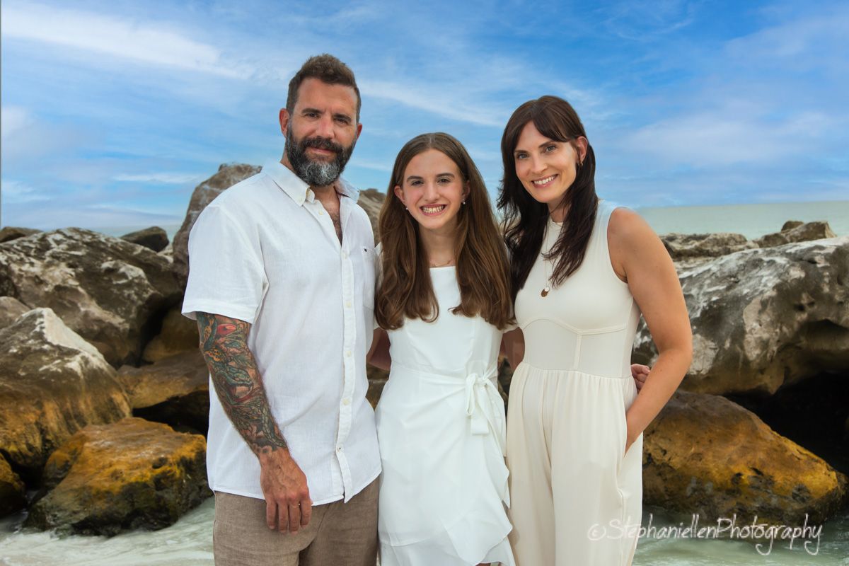 Family of three posing on rocky beach, man with tattoos in white shirt, two women in white dresses, blue sky backdrop.