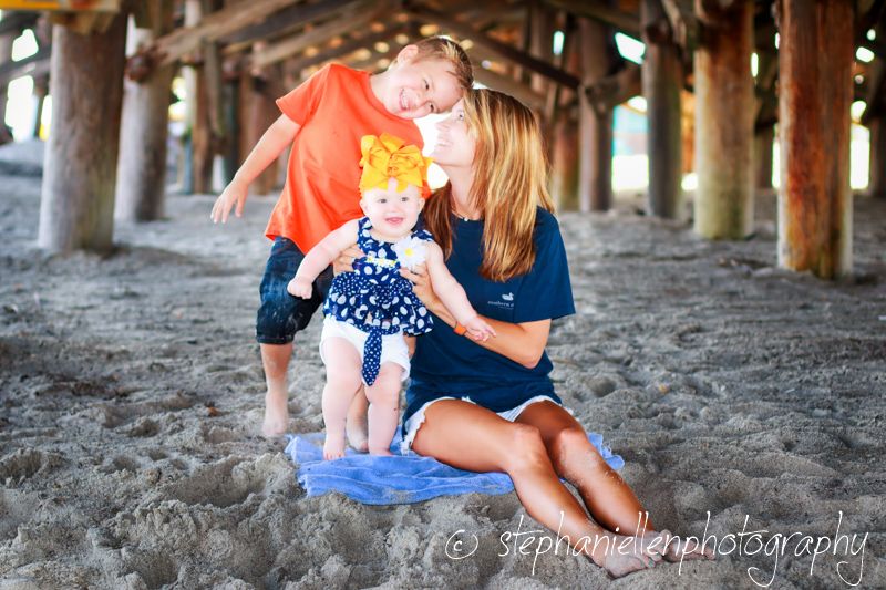 Mother sitting on beach sand under pier with two young children, family portrait photography.