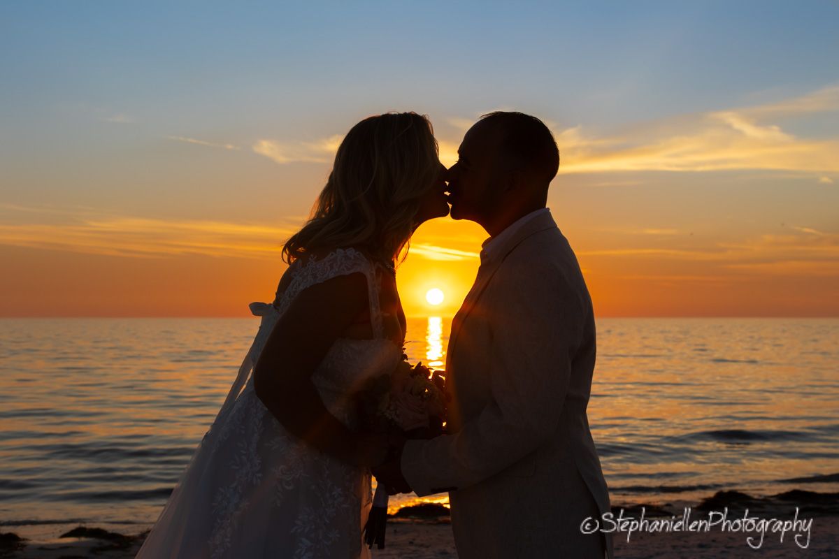 Bride and groom silhouetted kissing against a vibrant ocean sunset.