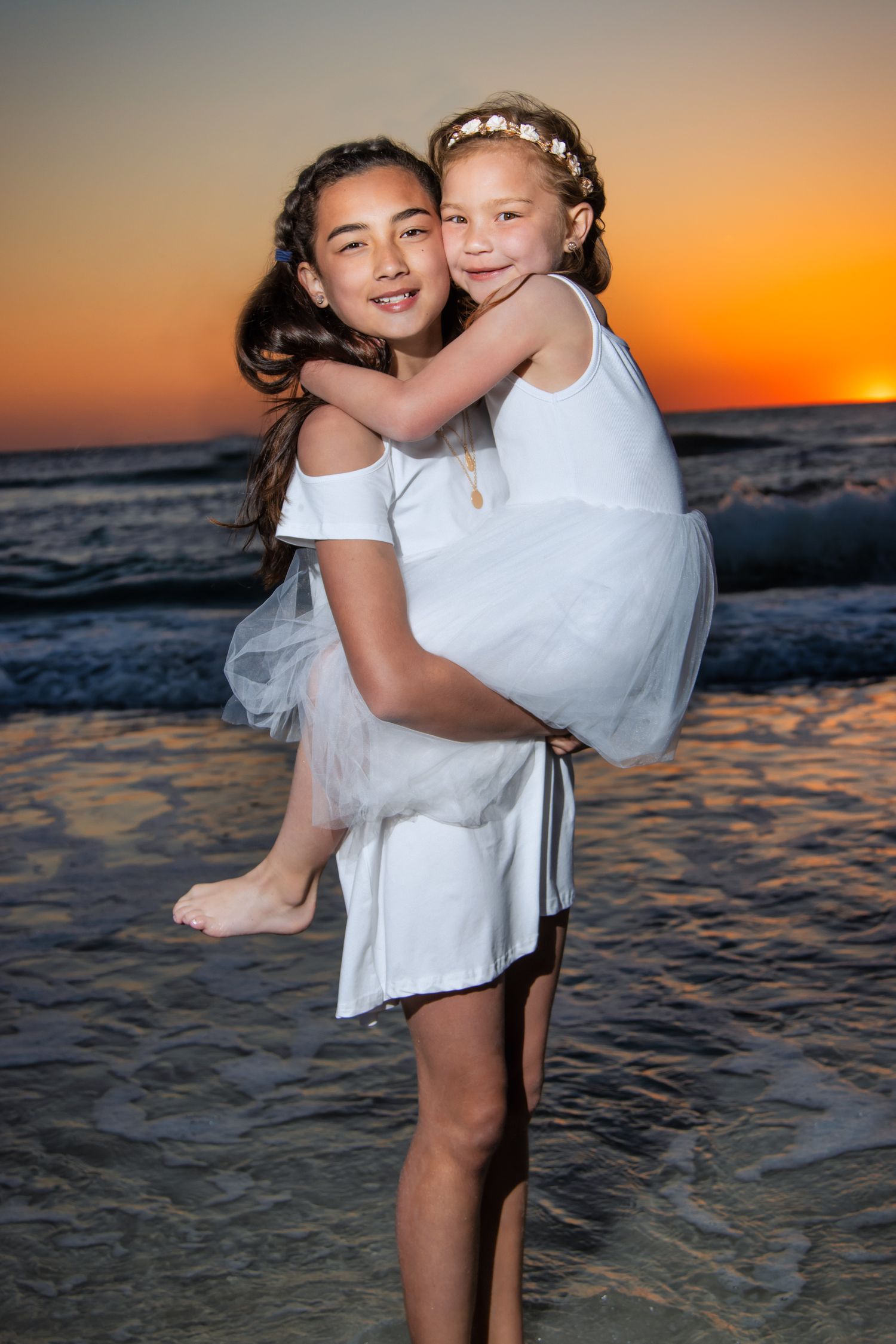Two girls in white dresses embrace on a beach at sunset, waves gently lapping the shore behind them.