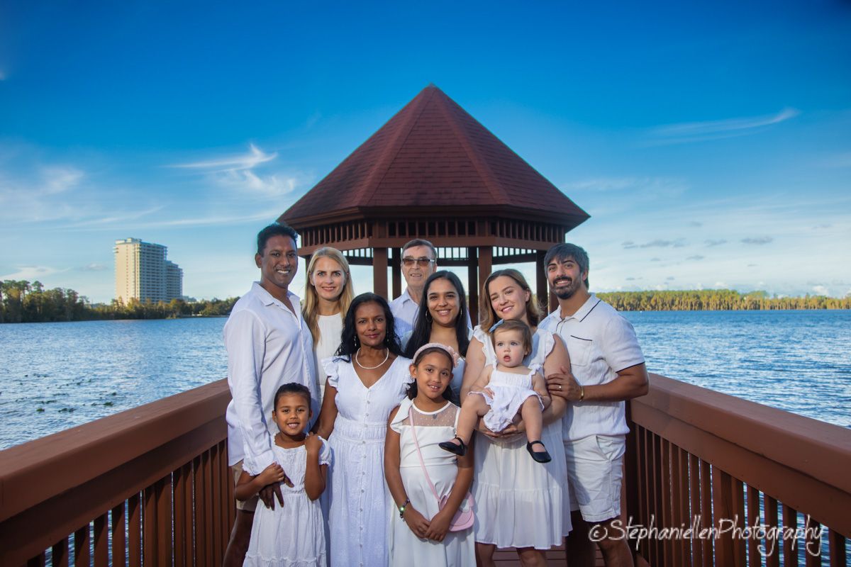 Family of nine in matching white outfits posing on a wooden dock with a gazebo and lake view.