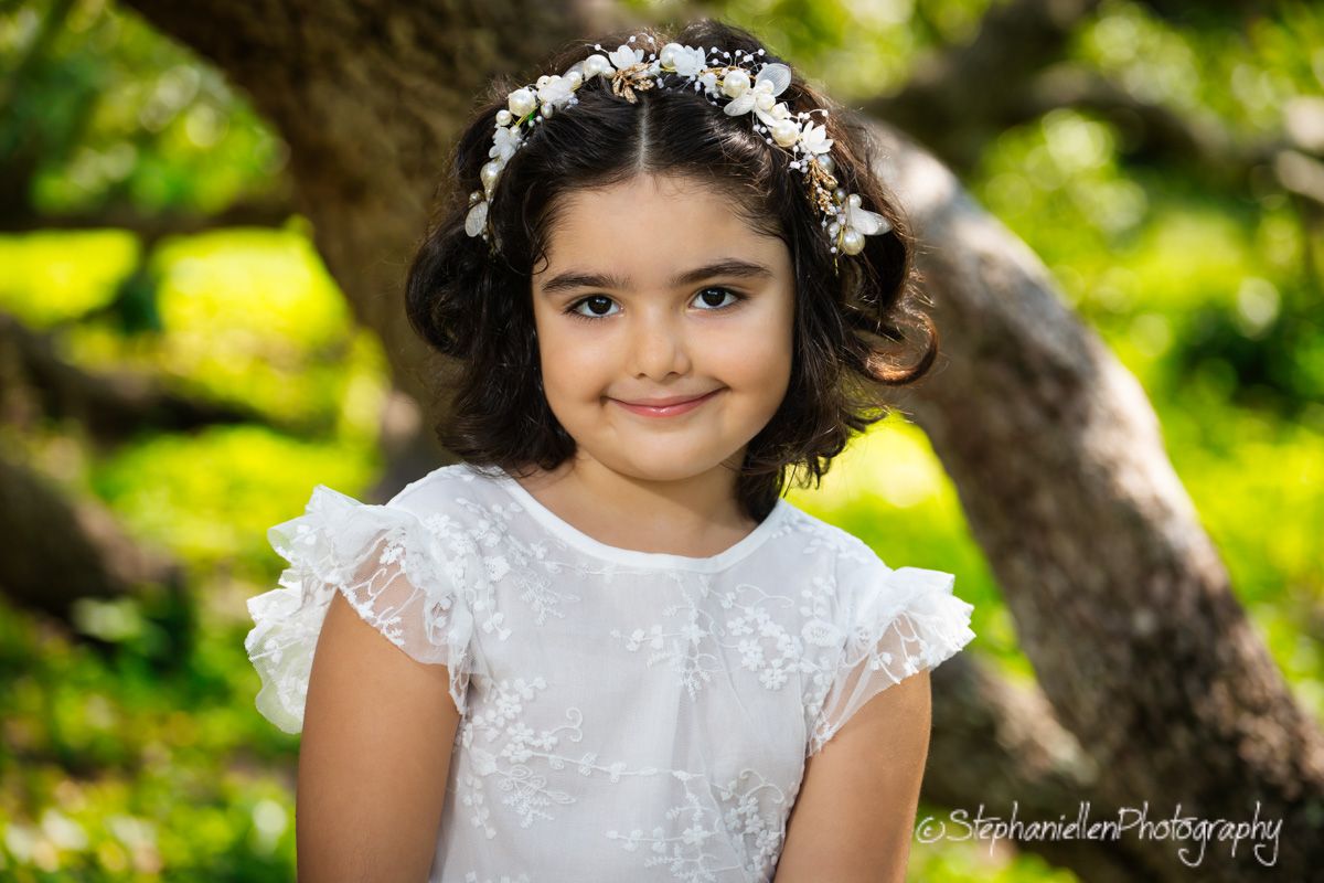 Young girl in white lace dress and floral crown smiles outdoors amid lush green bokeh background.