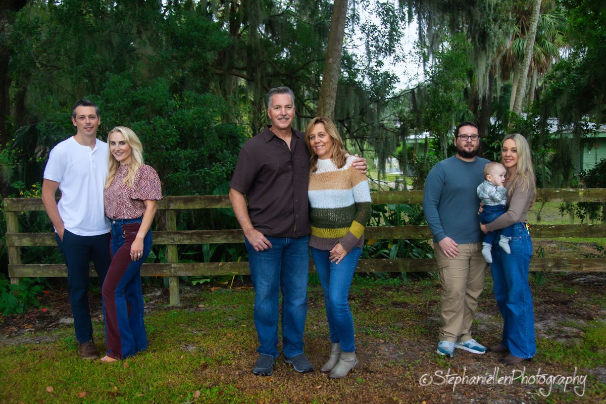Six people posing outdoors near a wooden fence surrounded by lush green trees in a family portrait photo.