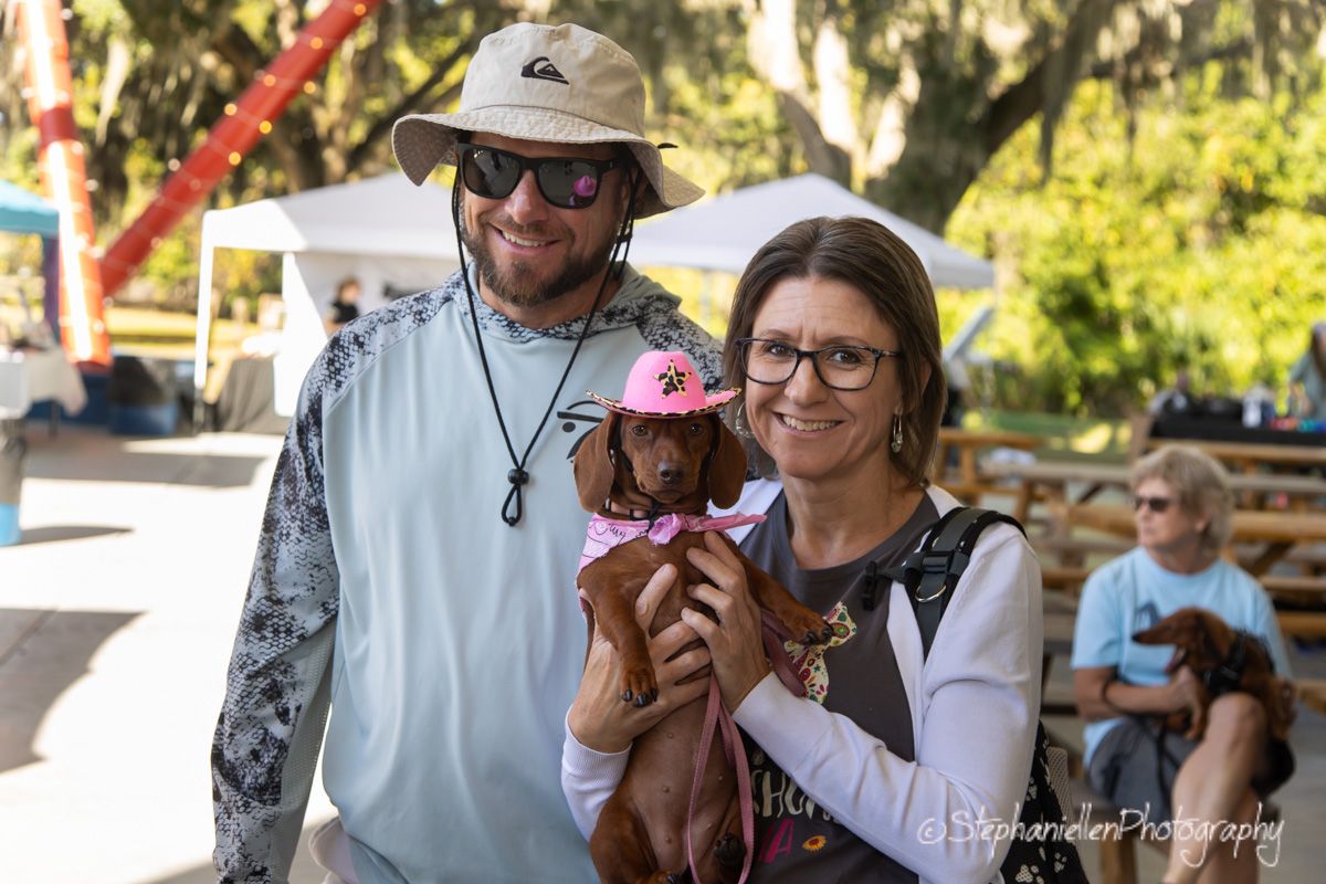 Couple smiling outdoors holding a dachshund in a pink outfit at a sunny park event.