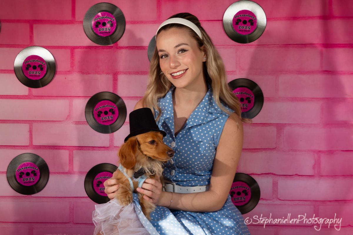 Young woman in blue polka dot dress holds a small dog wearing a top hat, posing with vinyl records on pink backdrop.