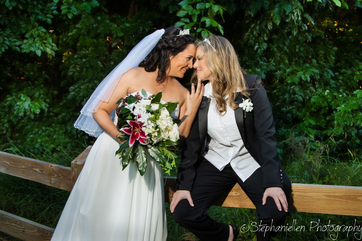 Two brides share intimate moment on fence, one in white gown with veil holding floral bouquet, other in dark suit.
