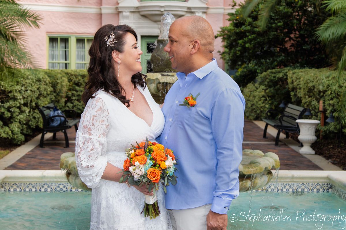 Bride and groom sharing a loving gaze holding orange bouquet by a pink building fountain.