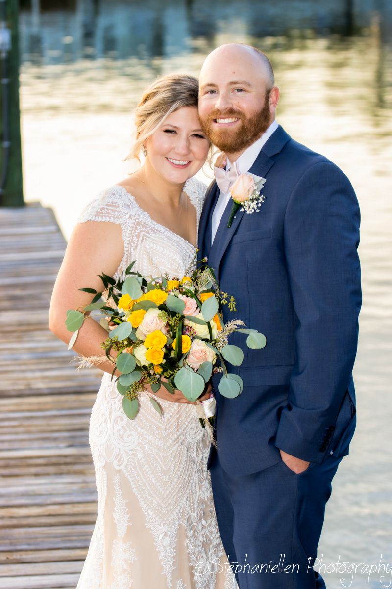 Smiling bride and groom pose at waterfront dock, she holds yellow and pink floral bouquet.