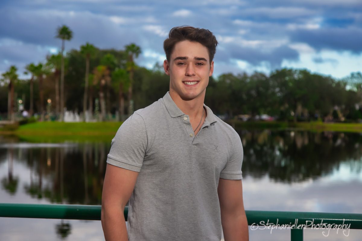Young man in gray polo shirt smiling outdoors near a pond with palm trees and cloudy sky backdrop.