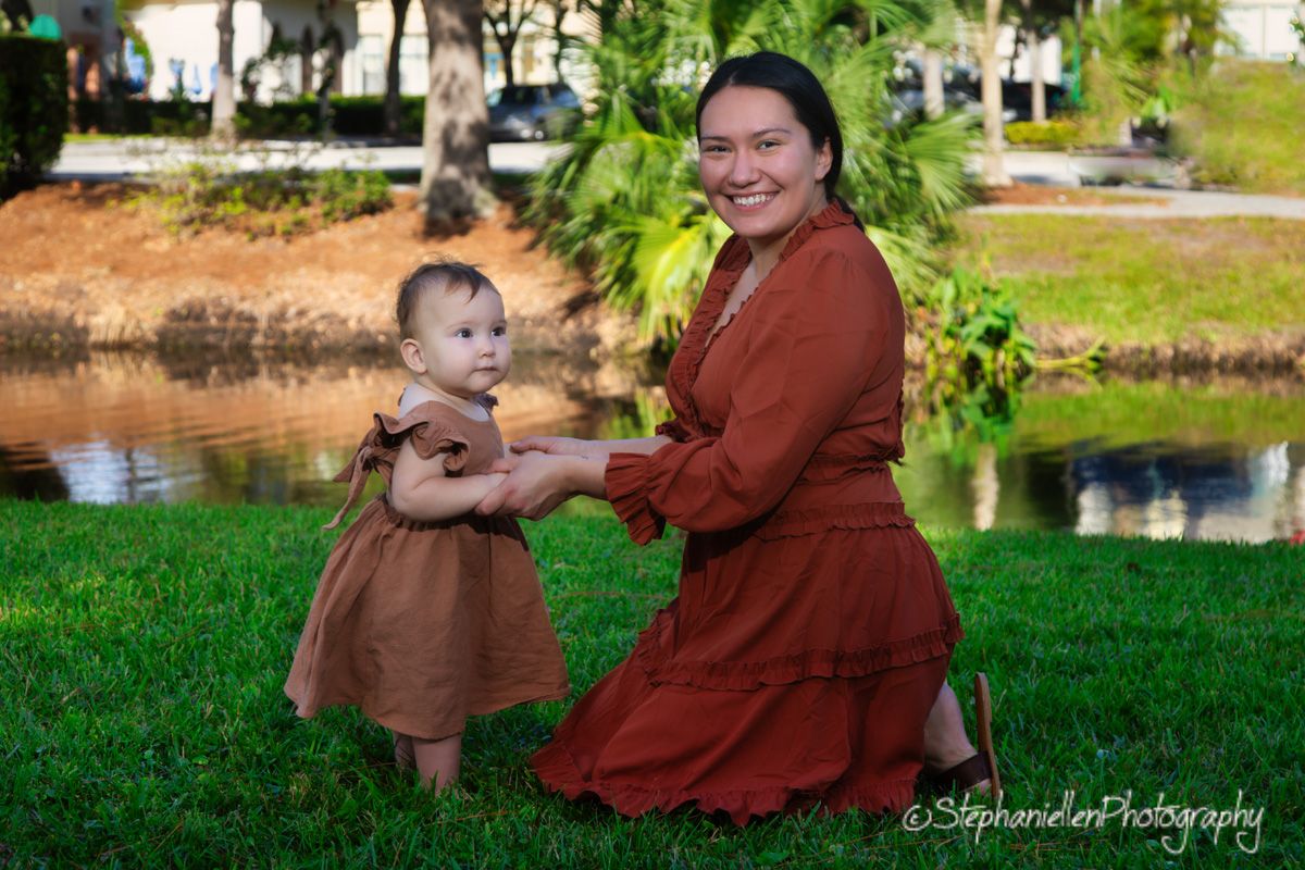 A mother kneels on green grass holding her baby girl's hands near a pond, both wearing matching brown dresses.