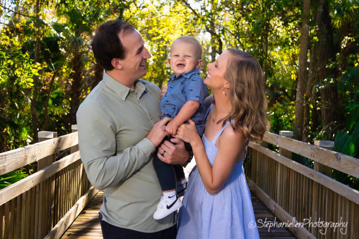 Happy couple holding smiling baby on a wooden bridge surrounded by green foliage.