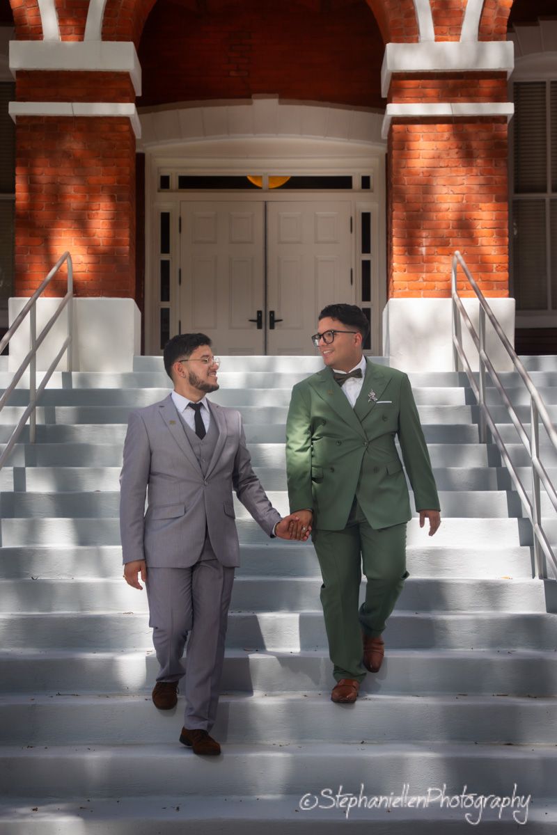 Two grooms in gray and green suits hold hands descending steps of a brick building.