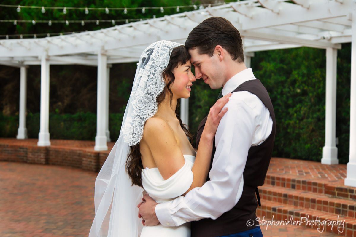 Bride and groom share tender moment under pergola with string lights.