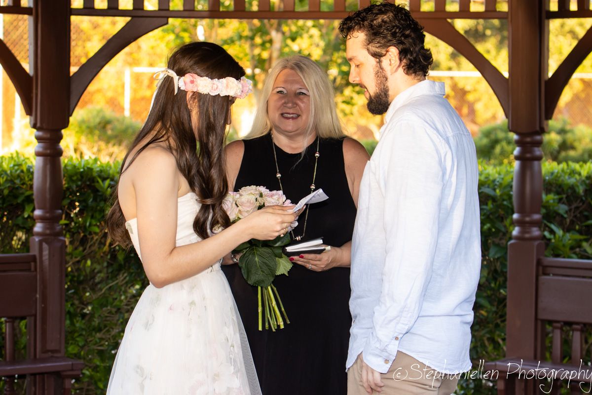Bride and groom exchange vows before a smiling officiant under a wooden gazebo at an outdoor wedding.