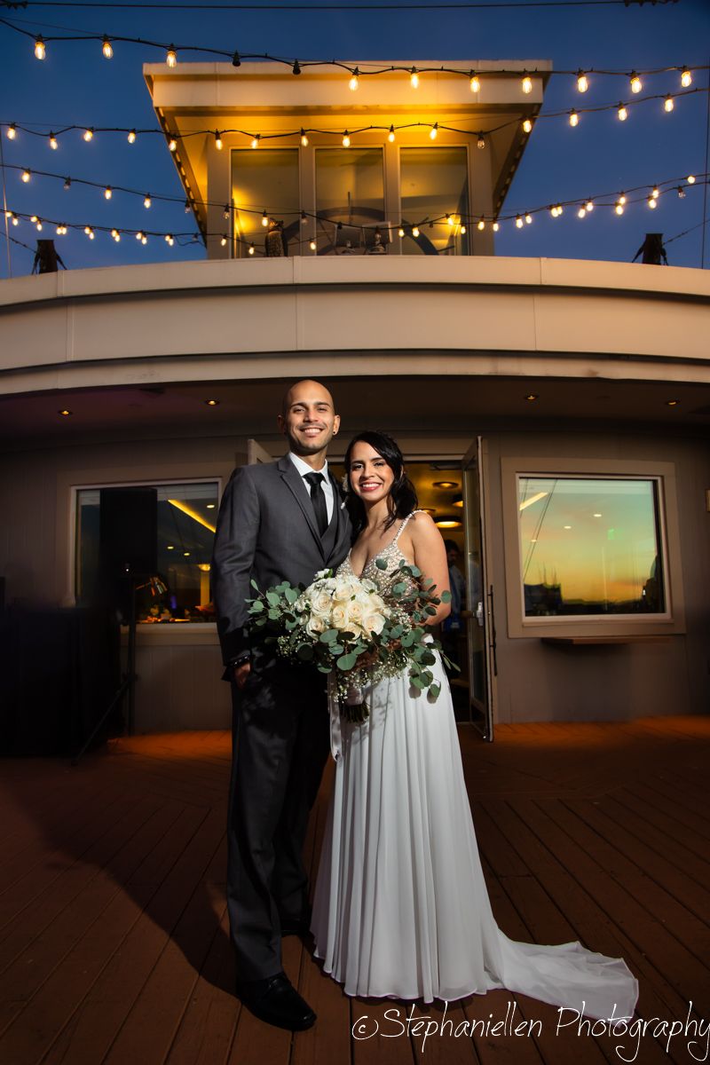 Bride and groom posing on a yacht deck at night with string lights glowing above them.