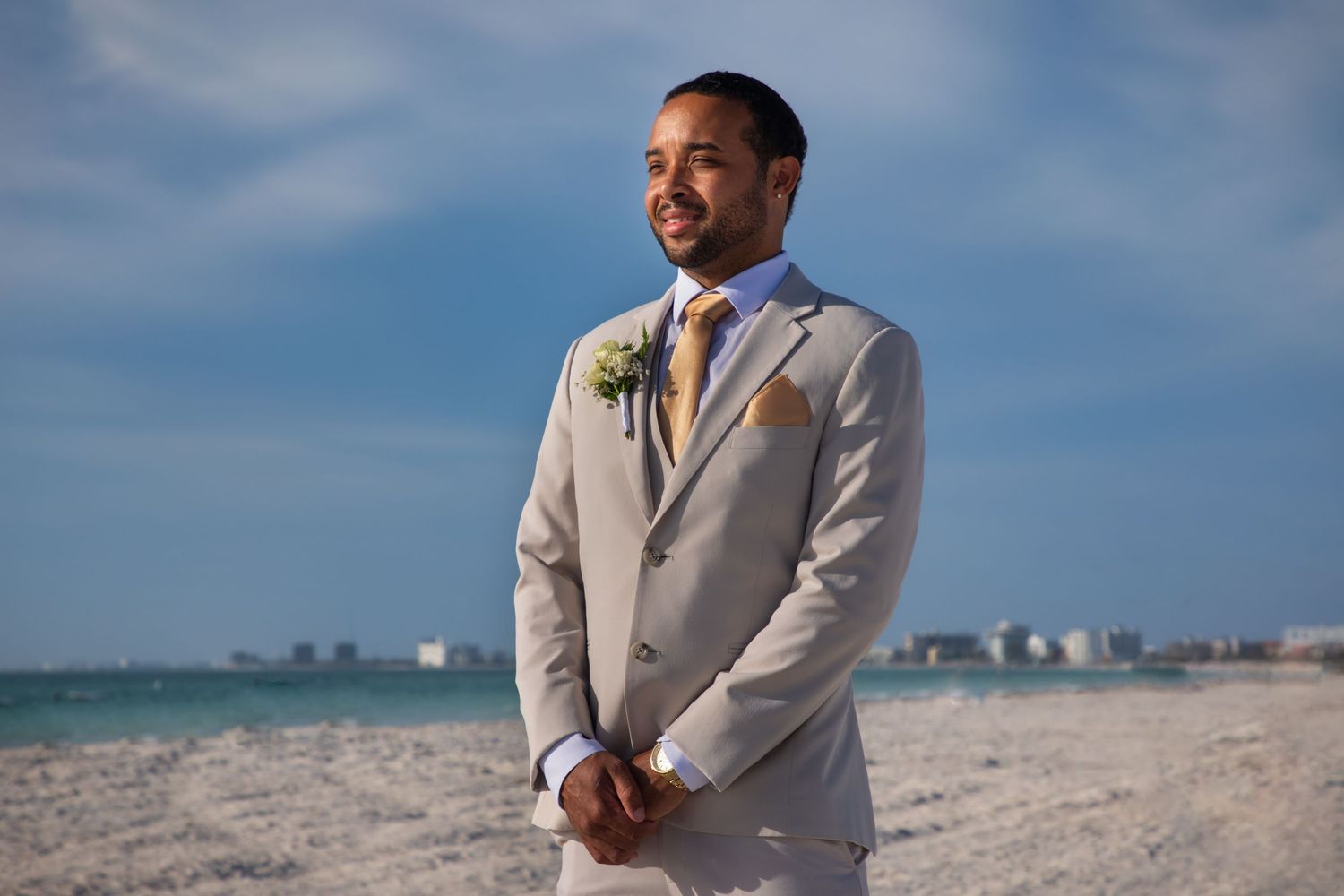 Handsome groom in tan suit with boutonniere standing on sandy beach under blue sky.