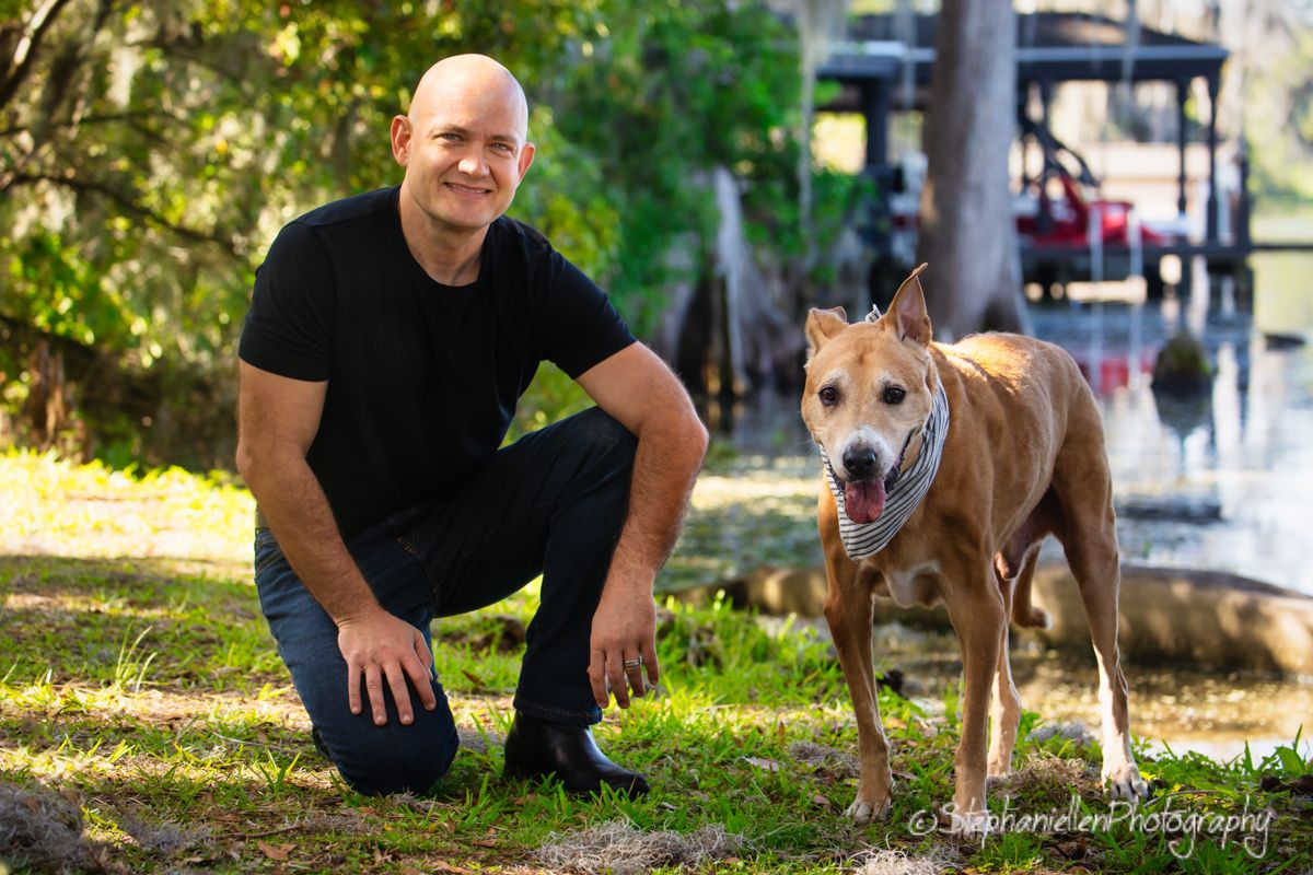 Bald man in black shirt crouching beside a tan mixed breed dog outdoors near a waterway.