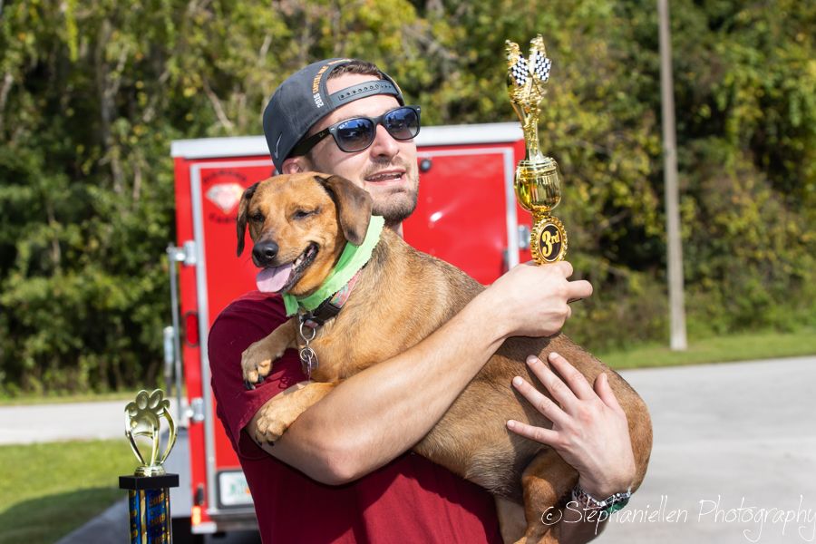 Smiling man in maroon shirt holds a happy dachshund and a trophy at an outdoor dog racing event.