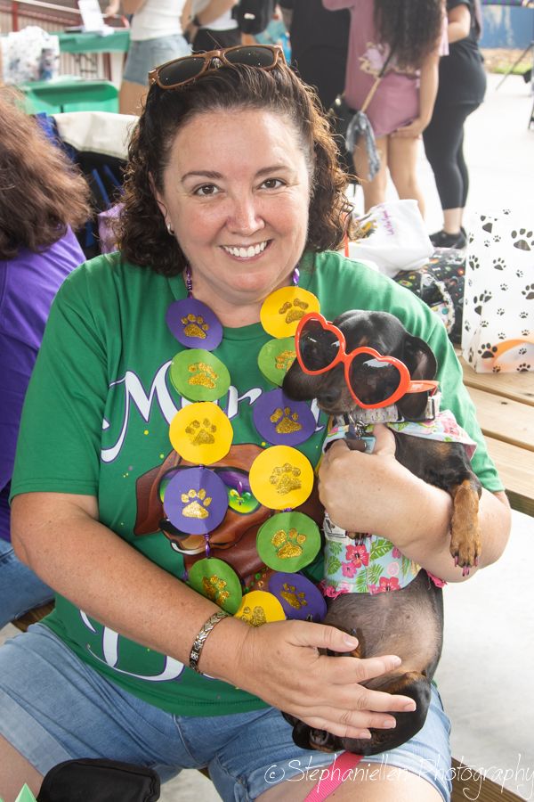 Woman in green shirt holds a dachshund wearing heart sunglasses and floral outfit, adorned with colorful paw print medals.