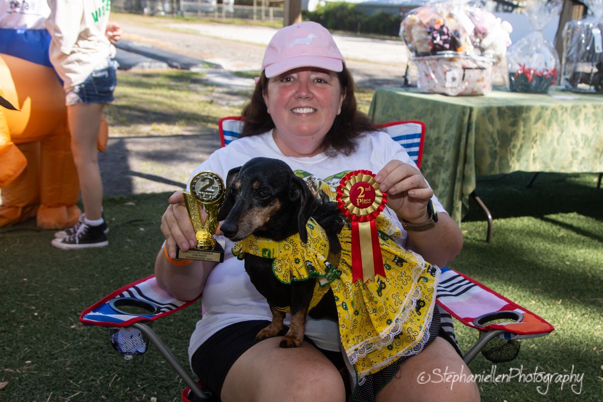 Woman in pink cap holds trophy and ribbon with dressed dachshund at outdoor dog show competition.