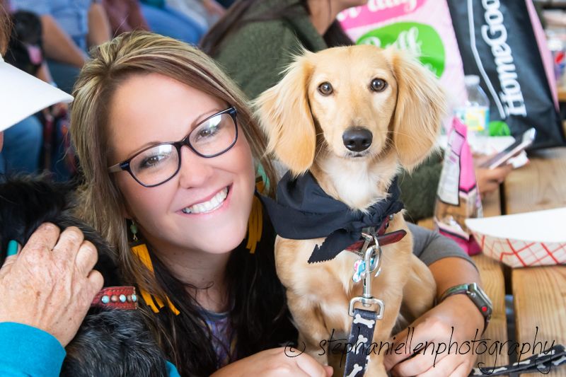 Smiling woman with glasses holds a cute long-haired dachshund wearing a black outfit at an outdoor event.