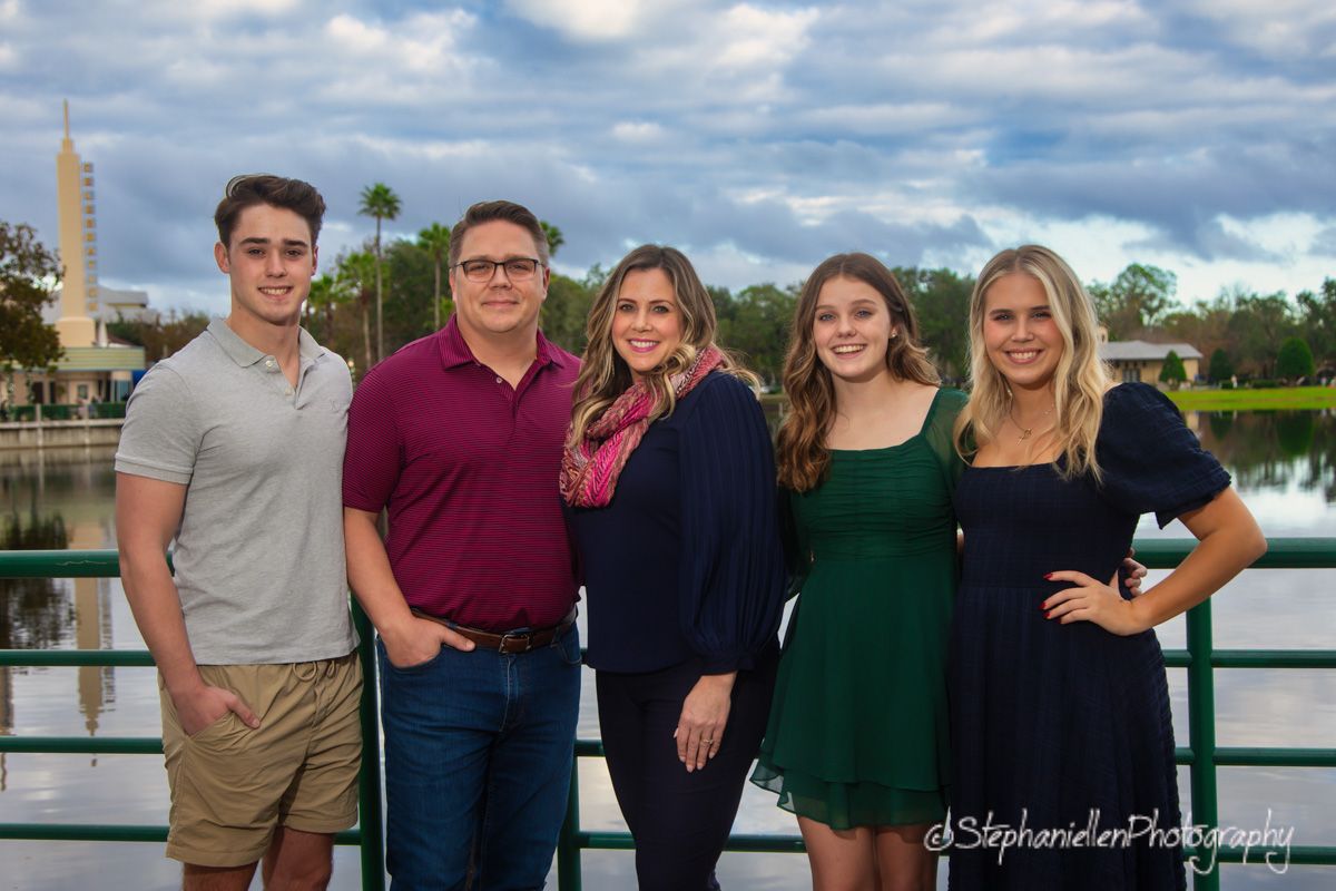 Family of five posing together outdoors near a scenic lake with cloudy skies and a tower in the background.