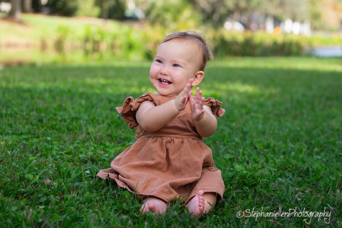 Smiling baby girl in brown dress sitting on green grass clapping hands during outdoor photo session.
