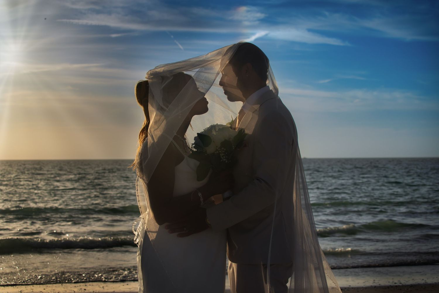 Bride and groom sharing intimate moment under flowing veil on beach at sunset.