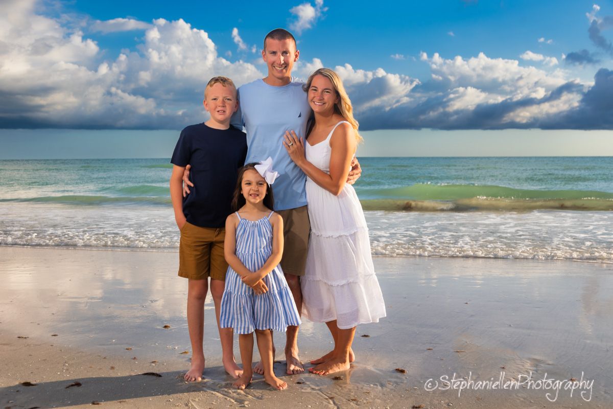 Family of four posing on sandy beach with turquoise ocean and dramatic cloudy sky.