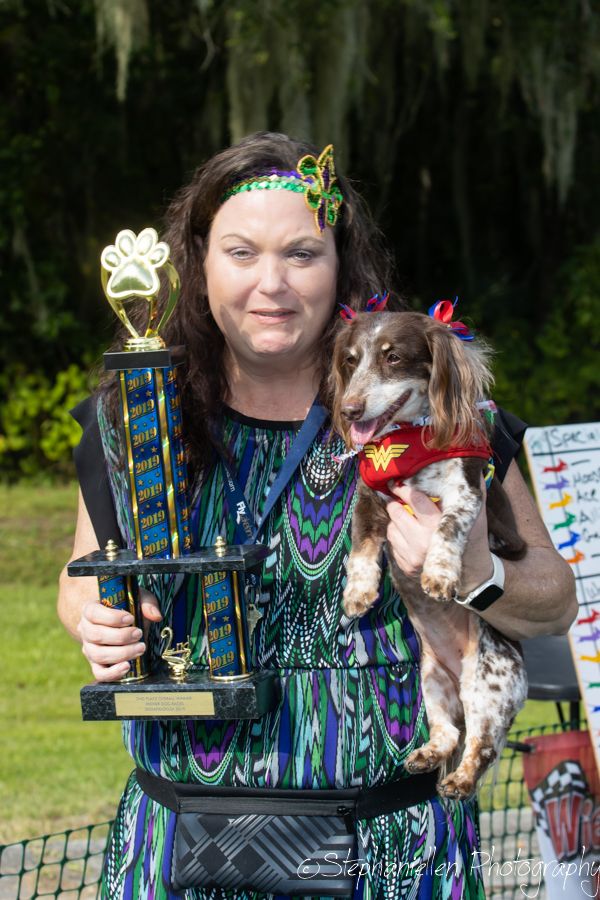 Woman holding trophy and costumed dachshund dressed as Wonder Woman at outdoor dog show competition.