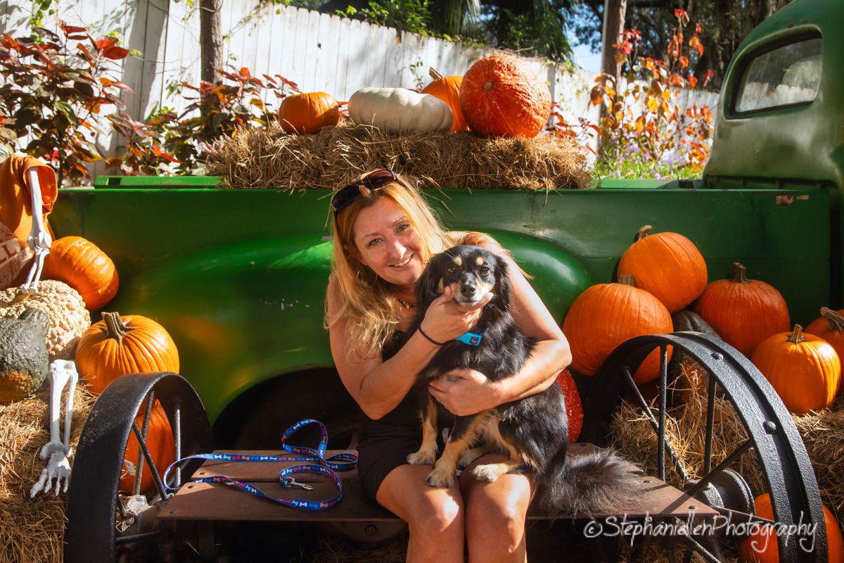 Smiling woman holding black dog on green vintage farm truck bed surrounded by orange pumpkins at Keel Farms Florida.