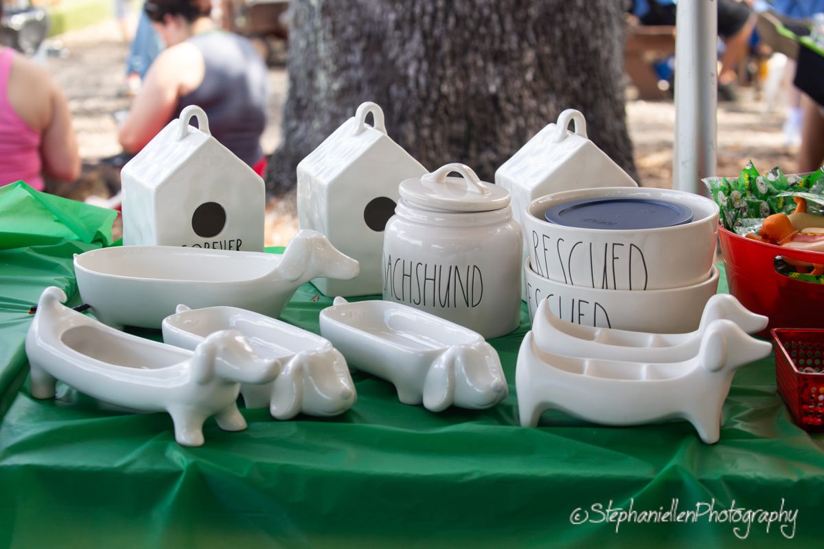 White ceramic dachshund dishes and birdhouses displayed on a green tablecloth at an outdoor market.