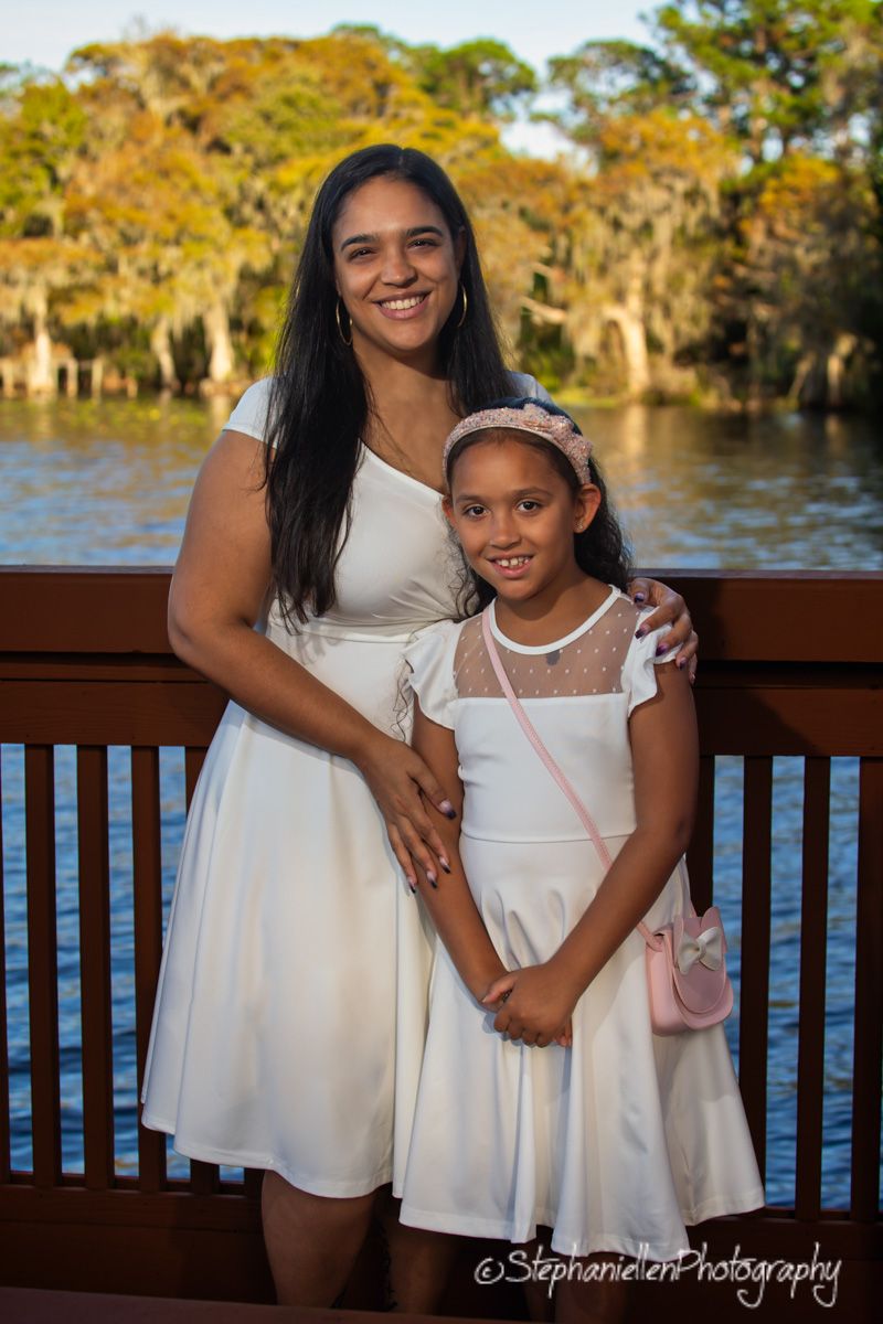 A woman and young girl in matching white dresses pose together on a wooden deck near a scenic waterway.