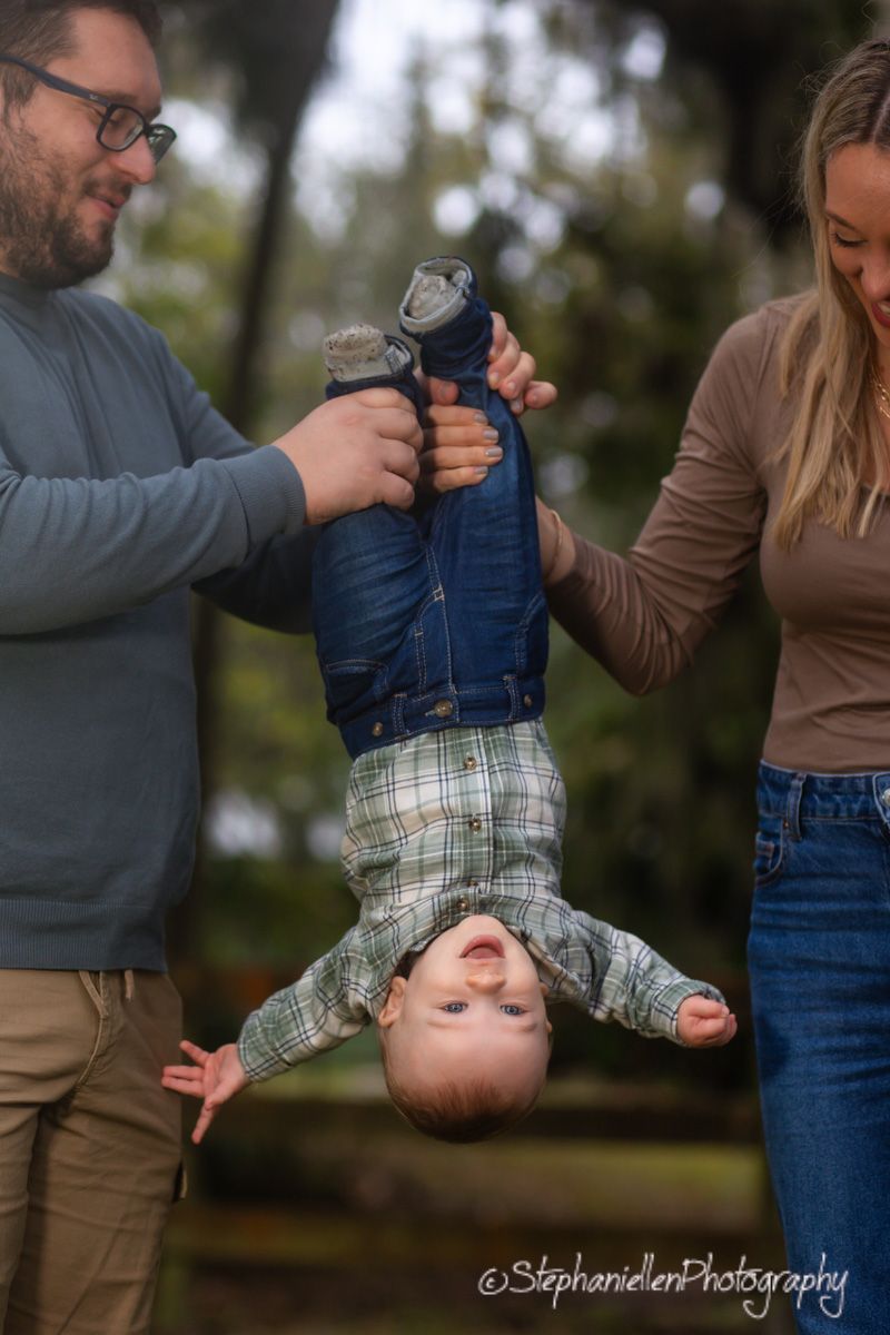 Parents playfully holding toddler boy upside down outdoors, all three laughing during family photo session.