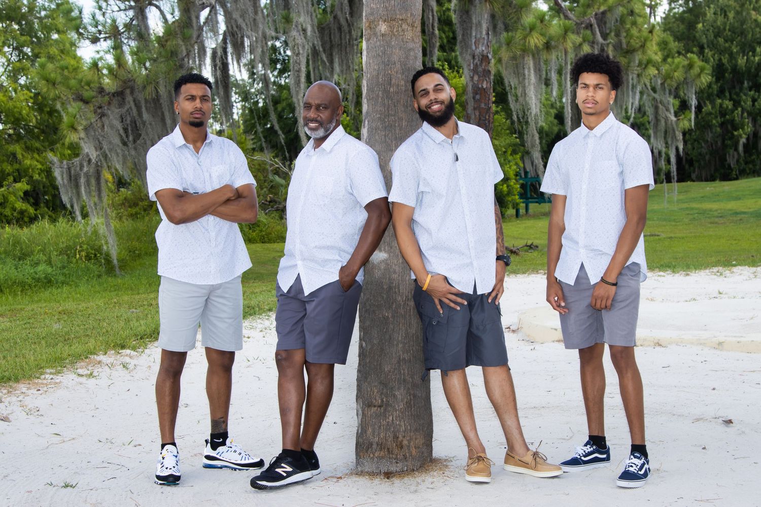 Four men in matching white shirts and gray shorts pose together near a palm tree on a sandy beach.