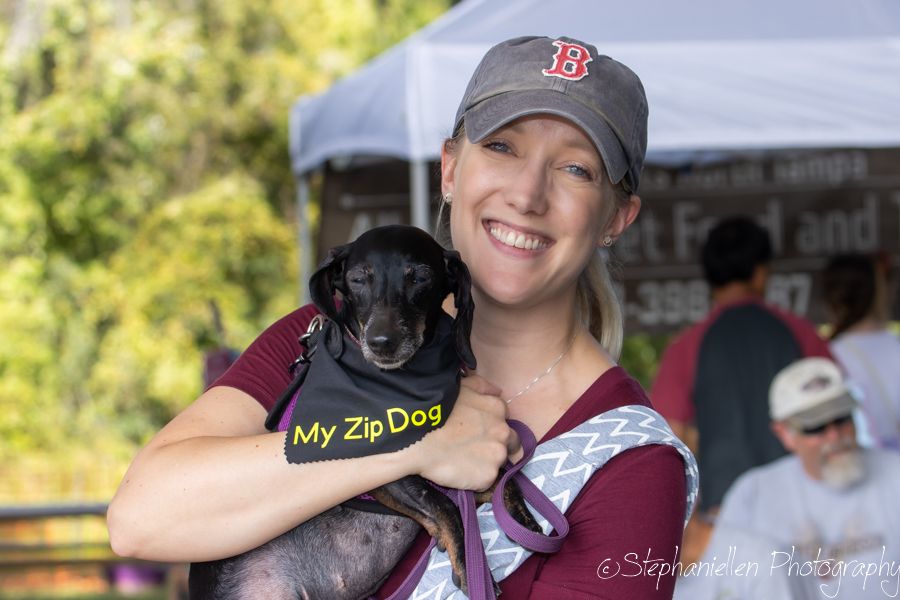 Smiling woman holding a small black dachshund wearing a yellow My Zip Dog vest at an outdoor event.