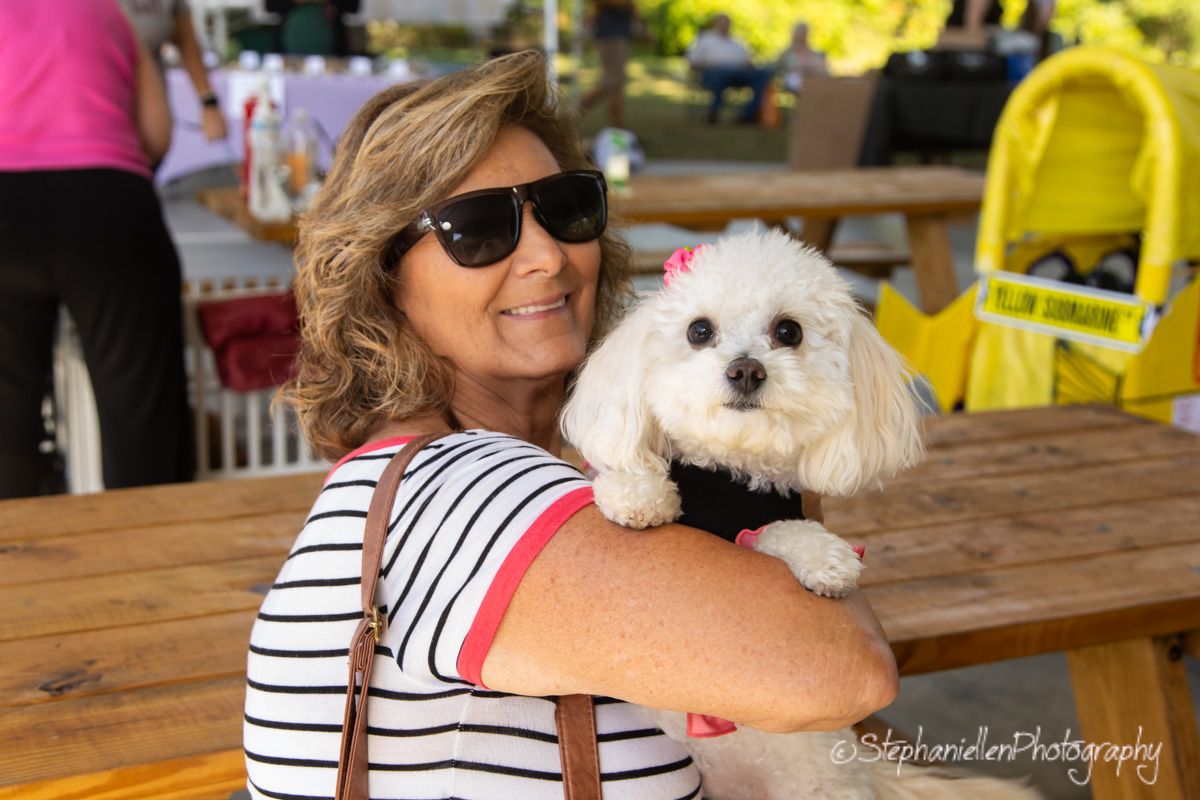 Woman in striped shirt and sunglasses holding a small white Maltese dog outdoors at a sunny event.