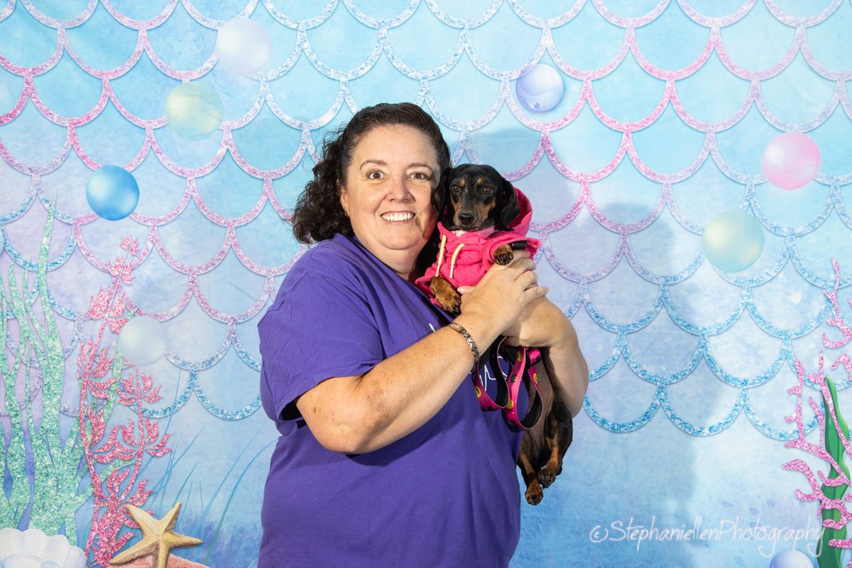 Woman in purple shirt holds a dachshund in pink outfit against a mermaid-themed backdrop.