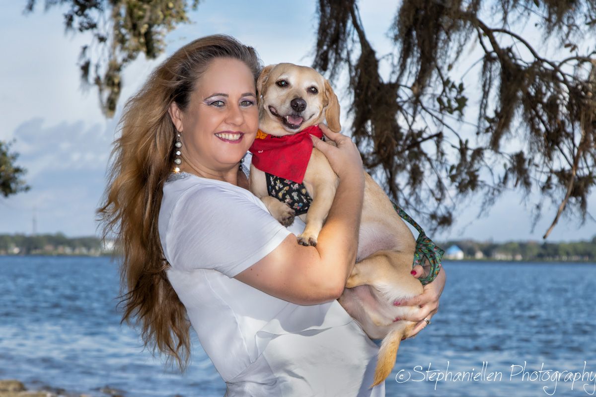 Smiling woman in white top holding a small tan Chihuahua in a red outfit near a waterfront.