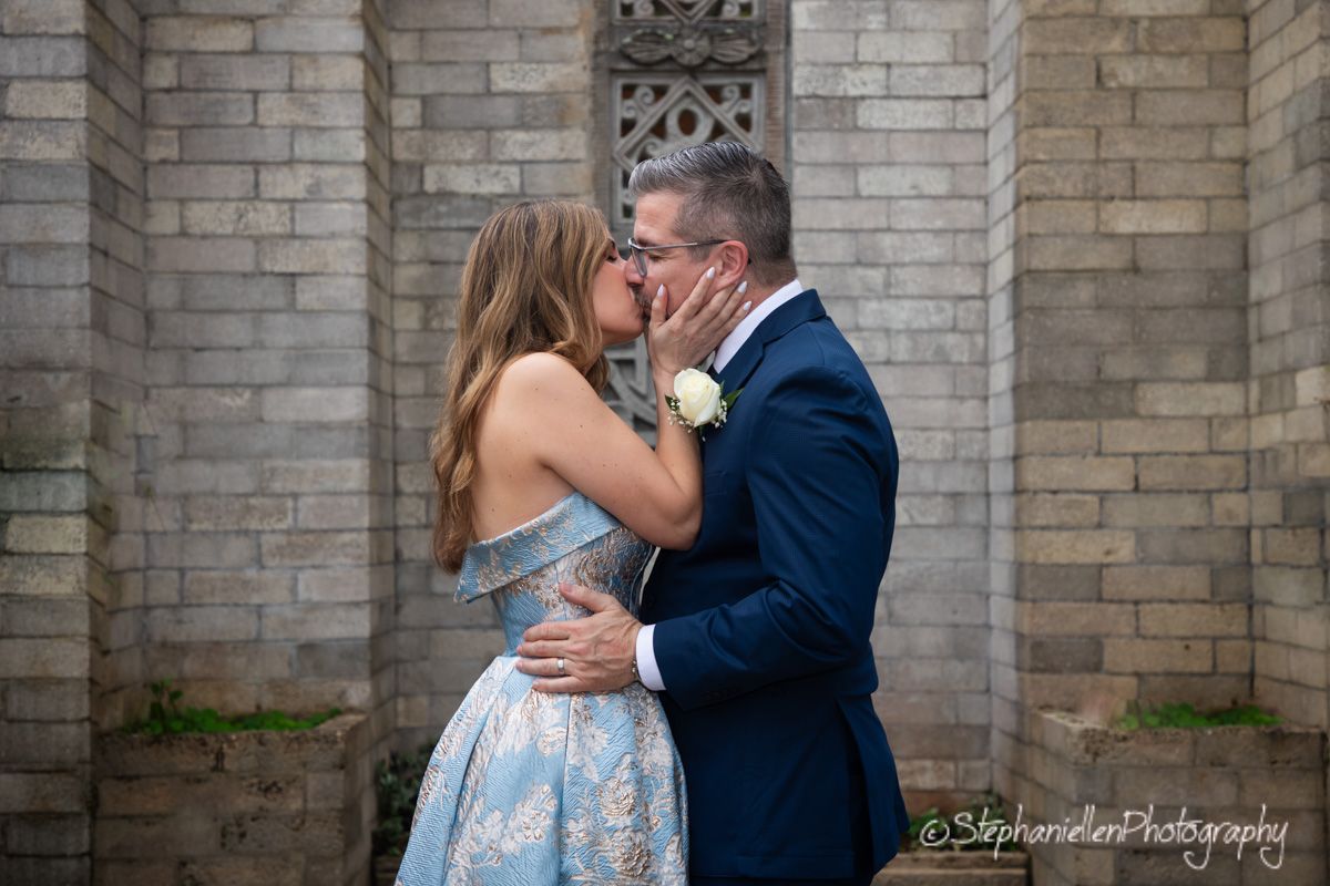 Couple shares a kiss in front of a stone chapel wall.