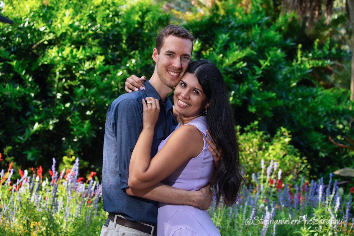 Smiling couple embracing in a colorful flower garden, woman in lavender dress hugging man in gray shirt.