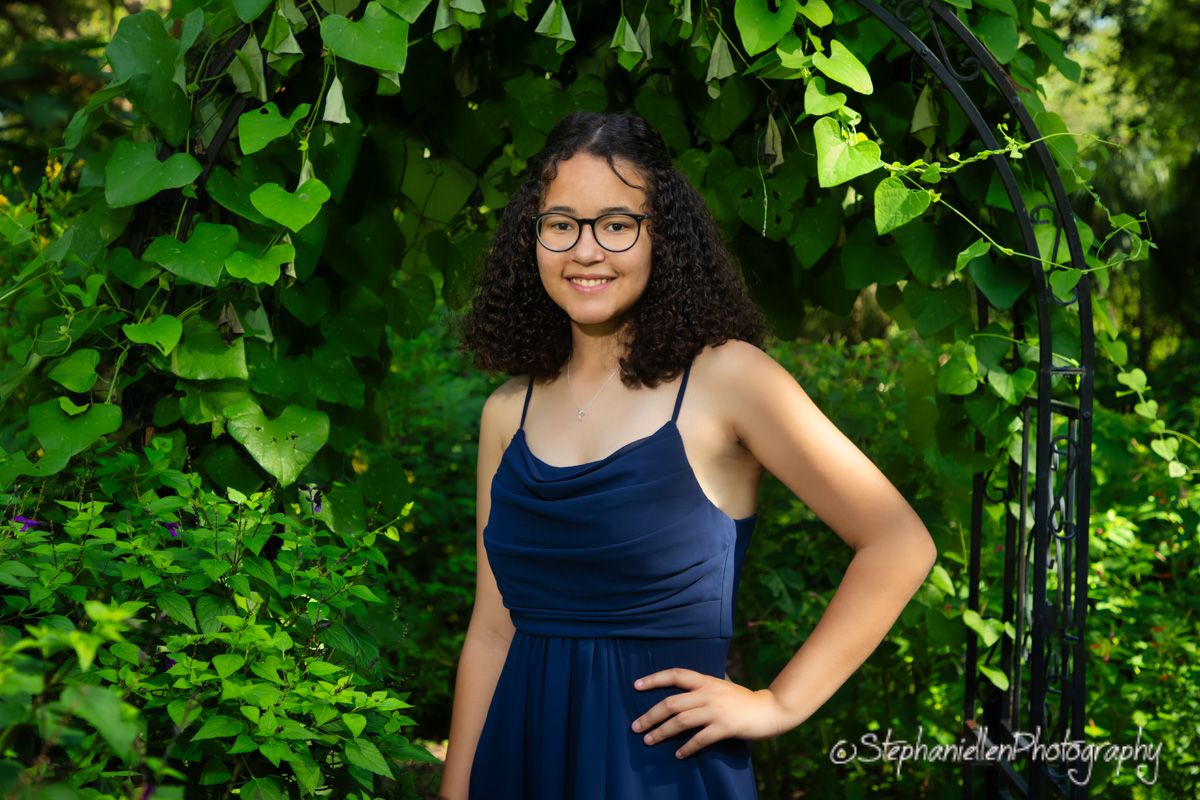 Young woman in navy blue dress standing under green vine archway outdoors.