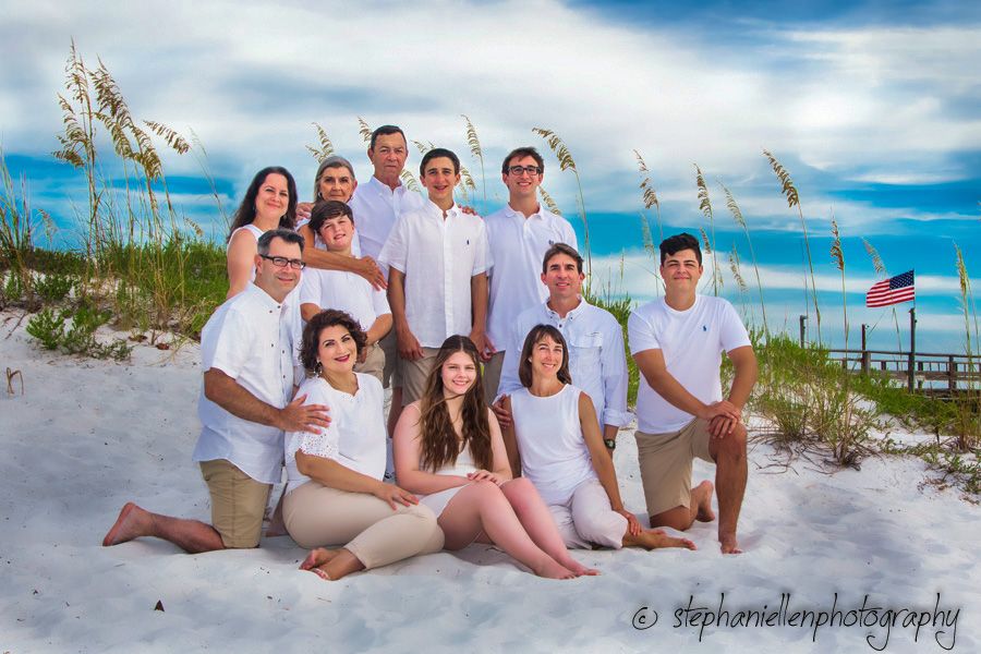 Family beach portrait in matching white shirts posed on white sand with sea oats and blue sky.