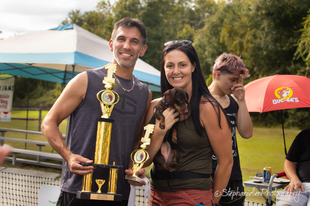 Smiling couple holding trophies and a dachshund dog at an outdoor pet racing competition event.